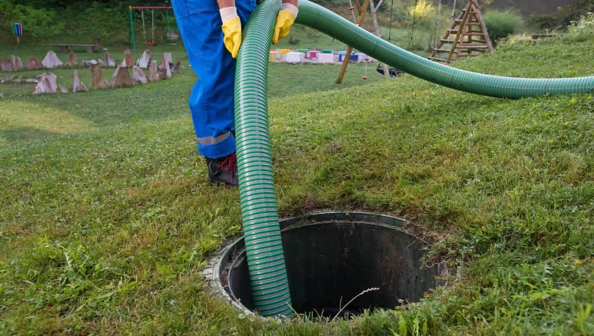 Technician in blue overalls pumping a septic system with a green hose on a grassy field.