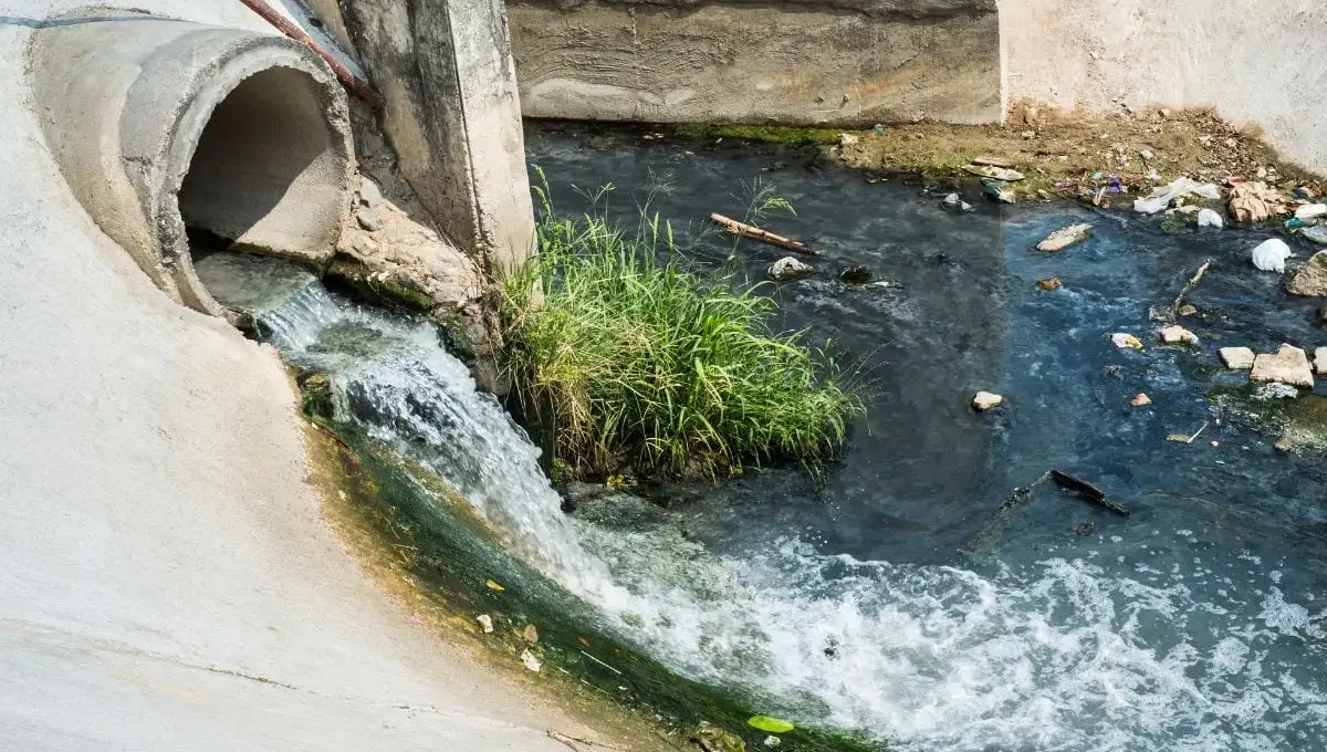 Wastewater gushing from a concrete pipe into a river, showing visible pollution and debris.